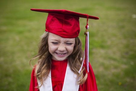 A cute little girl smiles happily in red cap and gown while standing on a field.