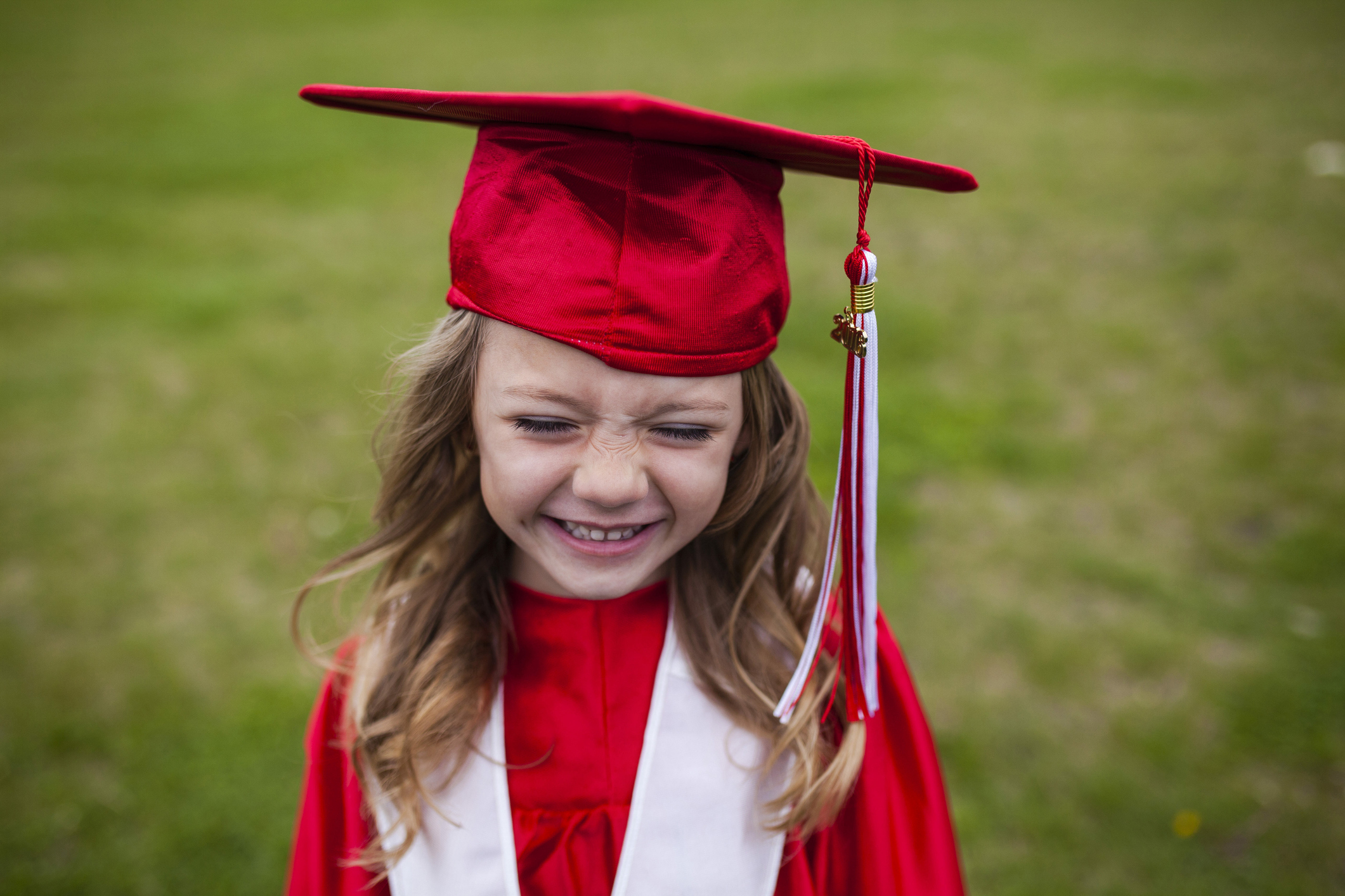 A cute little girl smiles happily in red cap and gown while standing on a field.