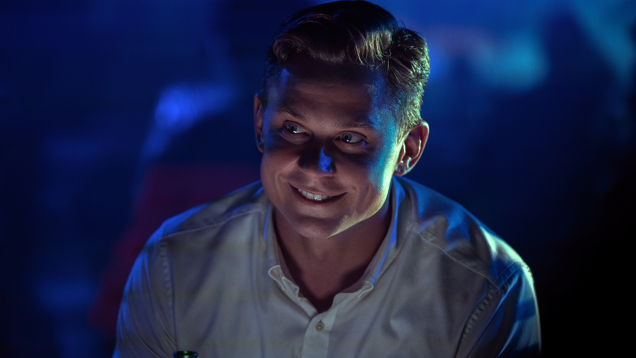 Billy Magnussen sitting in the club, smiling eerily, in No Time To Die.