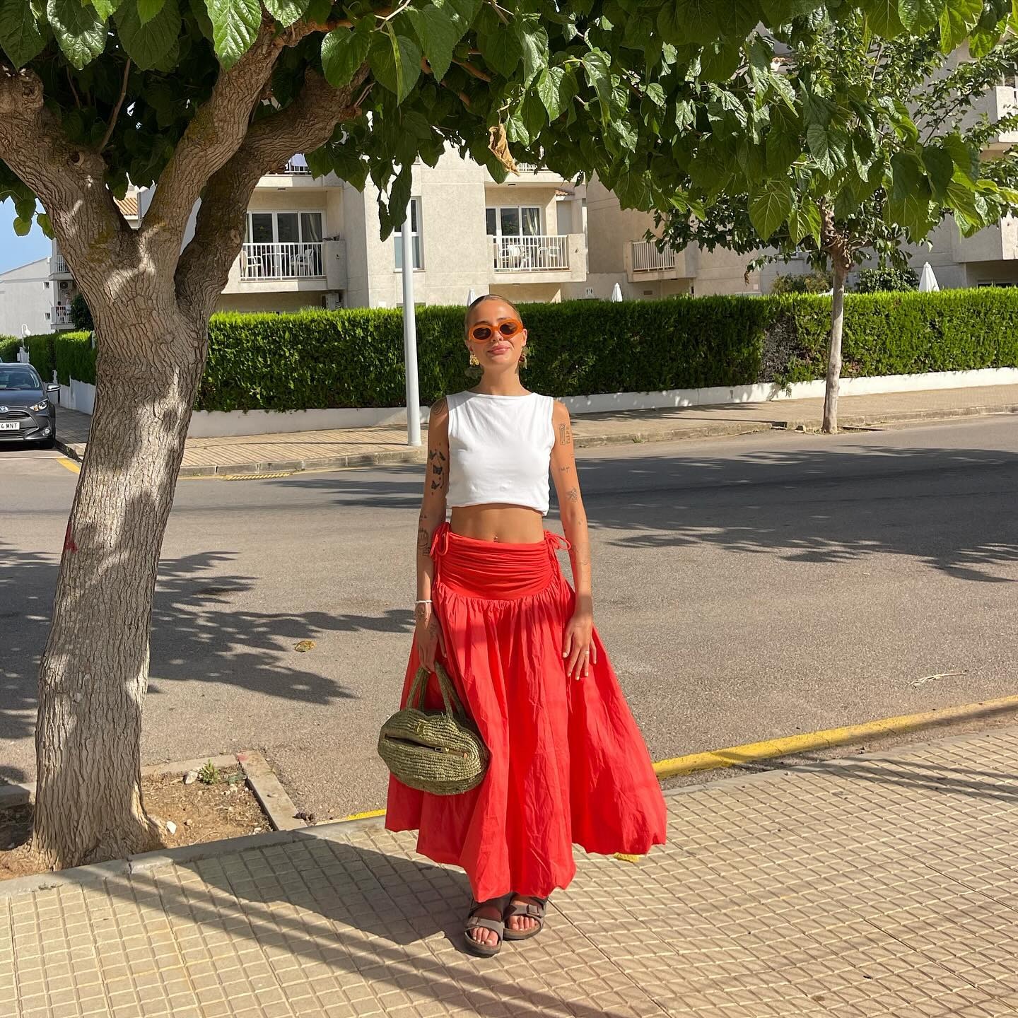 Fashion person Evie Mills has a red skirt outfit. She pairs her red skirt with a white cropped top, khaki green dad sandals and khaki raffia bag and orange ombree sunglasses. She is stood smiling at the camera in recent image taken from her Instagram.