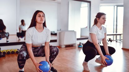 Two women in fitness studio holding blue Pilates ball squat down, the heels of the woman on the right are off the floor