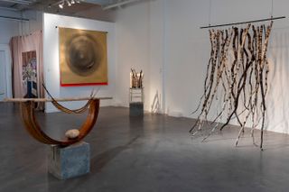 An exhibition room features a series of installations, including hanging branches in a swing-like structure, captured on the right, a wood, rock, and cement sculpture on the left, and bidimensional paintings at the back.