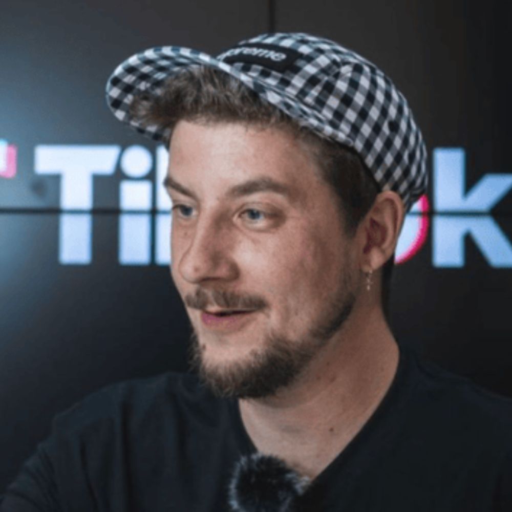 A man with a short beard and mustache, wearing a black T-shirt and a gingham-patterned cap. He has a small hoop earring in one ear and is positioned against a dark backdrop featuring blurred text and colourful graphics.