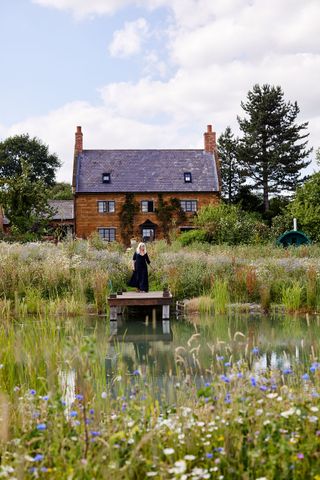 stone cottage with natural swimming pond with woman standing on jetty with wildflower meadow around