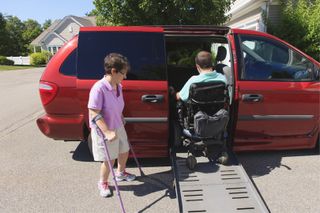 A man in a wheel chair goes up a ramp into a minivan as a woman using canes to walk stands by and watches.