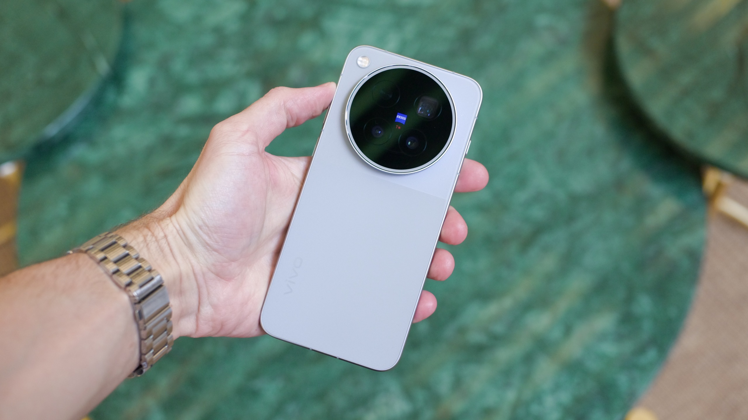 Vivo X500 Pro Max held in one hand, showing its silver back and large circular Zeiss camera module against a green marble background.