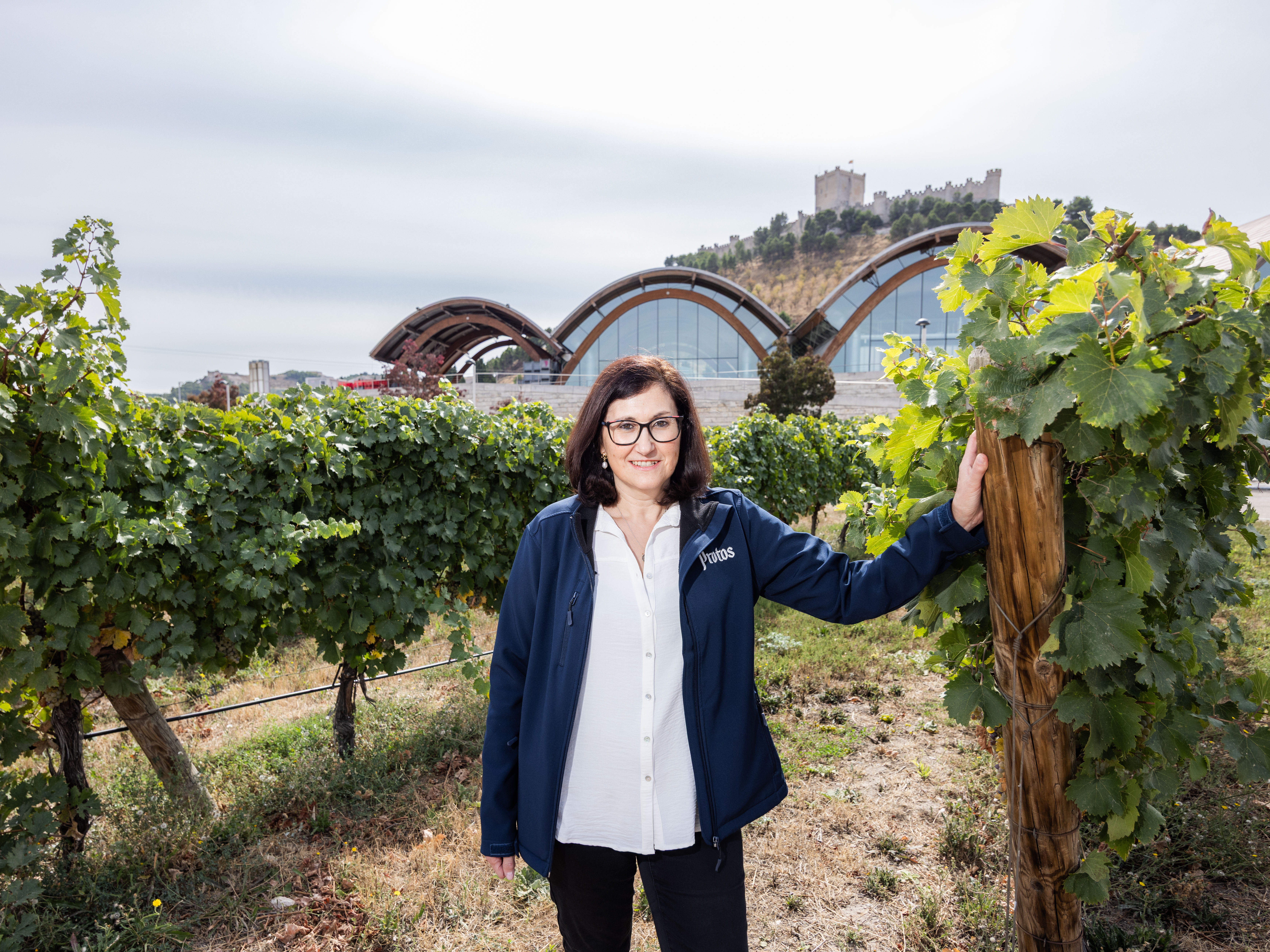 Marilena Bonilla standing between two rows of vines, with her hand up against a wooden post and the winery in the background