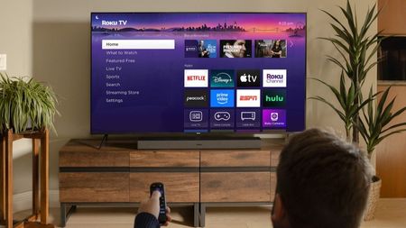 A man sat on a couch pointing a remote at a Roku TV 