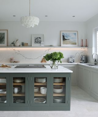 An L-shaped kitchen with a sage green kitchen island with a marble countertop, white cabinet and shelves with lighting behind it, and a white pendant light above it