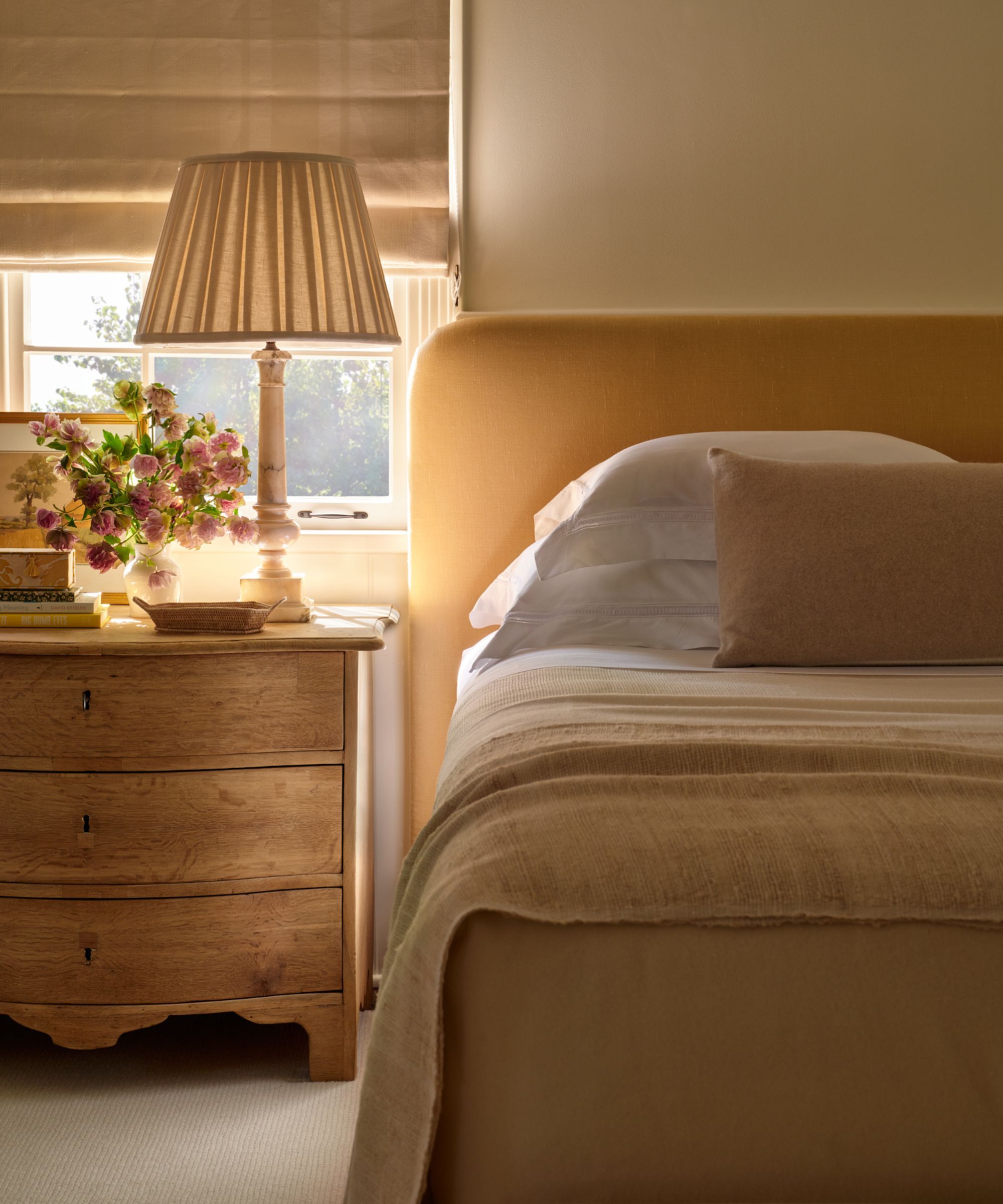 a cozy warm neutral bedroom with a yellow upholstered bed, simple neutral linens, wooden nightstand styled with a lamp and flowers