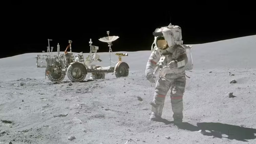 An astronaut walks on the moon with a rover behind them