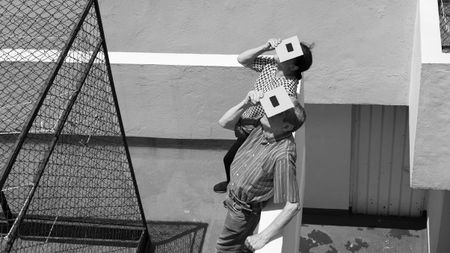 Two people, facing up, cover their heads with cardboard boxes having square cutouts. The setting seems outdoors with geometric shadows and lines