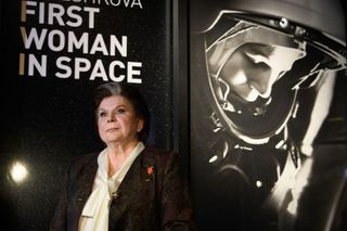 Valentina Tereshkova stands next to an image of herself from the Vostok 6 mission with the words "first woman in space" written in white font behind her.