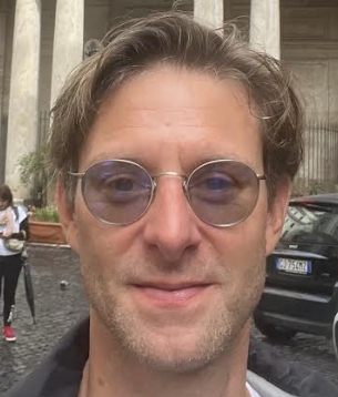 Headshot of a man wearing round sunglasses with dark blond hair