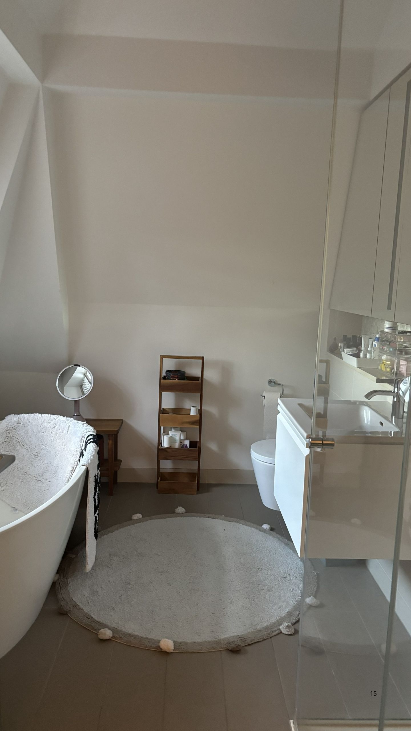 Image of a white and gray bathroom with a tub, a floating sink, a circular rug, a glass shower, and a wooden cady.