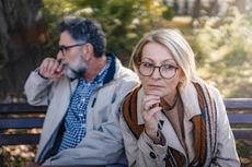 An older couple is sitting apart on a park bench in an autumn setting, looking upset and distant, portraying disagreement, relationship challenges, or emotional disconnect.