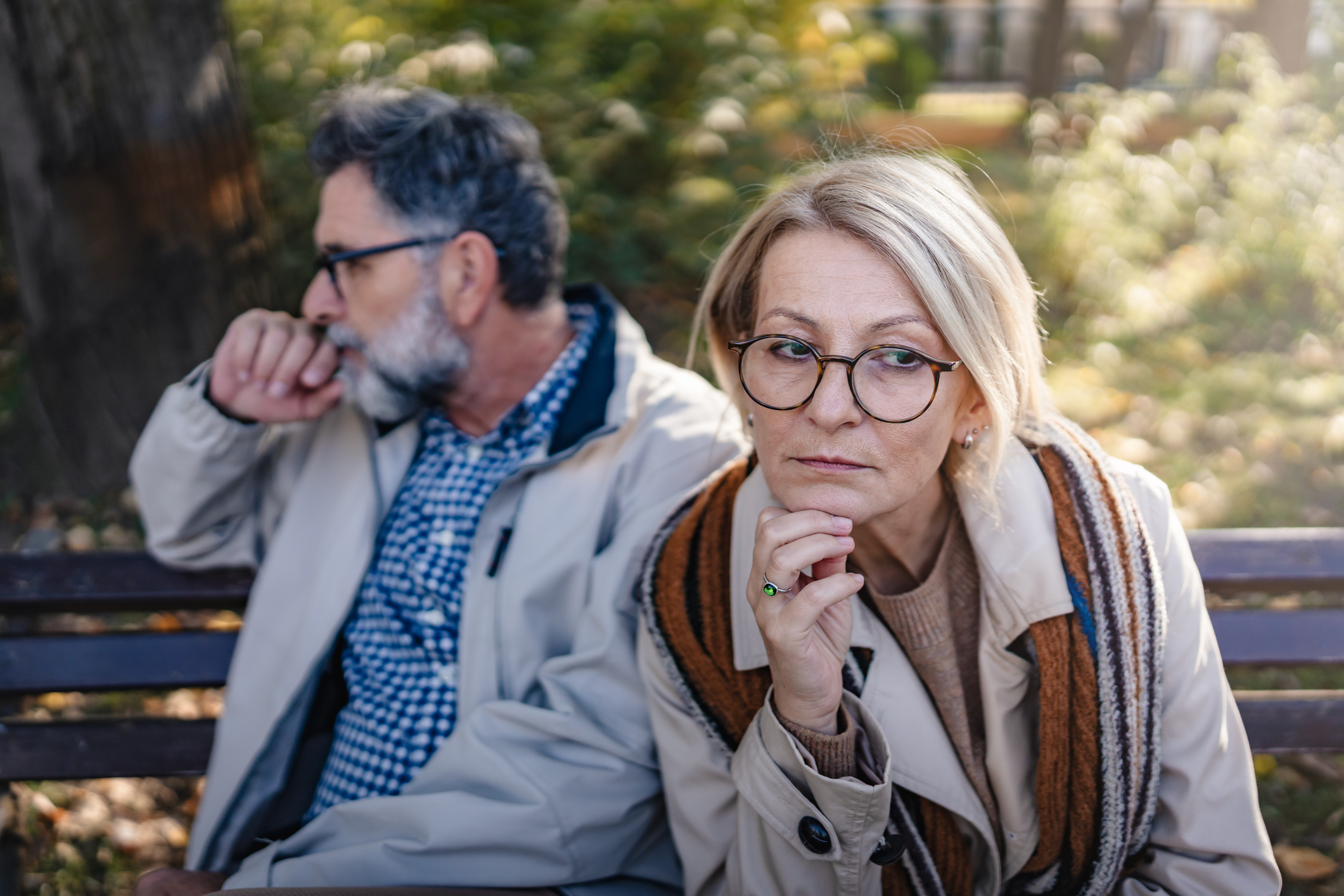 An older couple is sitting apart on a park bench in an autumn setting, looking upset and distant, portraying disagreement, relationship challenges, or emotional disconnect.