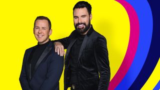 Scott Mills and Rylan stand in front of the 2023 Eurovision logo which shows a series of concentric hearts in different colours