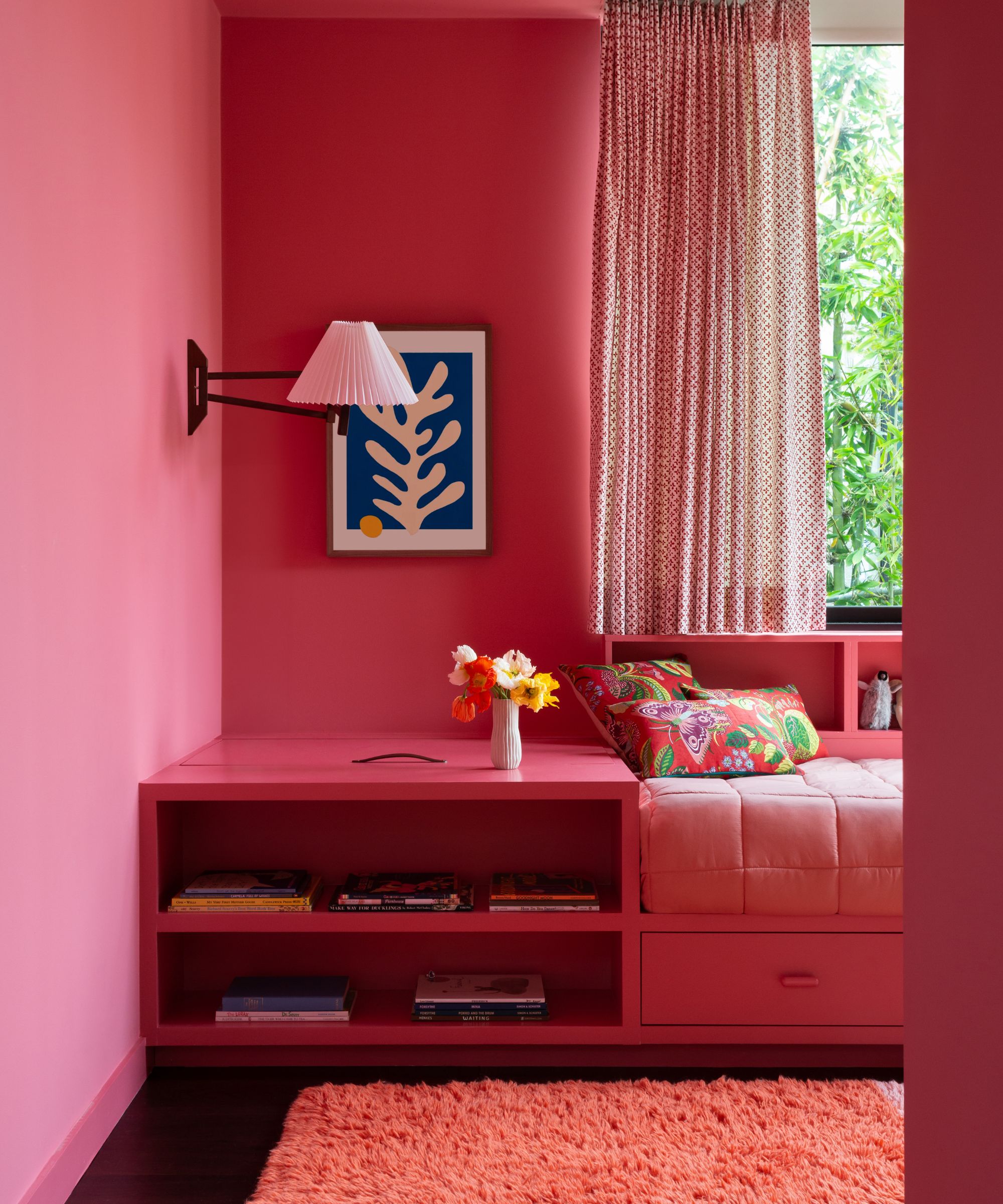 Girl's bedroom with a built-in bed and storage painted raspberry pink, a pleated sconce, a Matisse-style blue and white print, a large window with sheer polka dot curtains, and a fluffy pink rug.