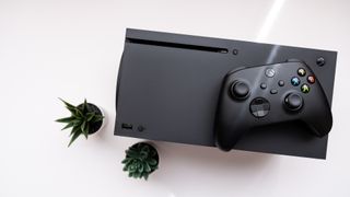 En Xbox Series X-konsol och en handkontroller ligger på ett vitt bord bredvid två små gröna växter.