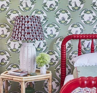 Green printed wallpaper behind a red painted bed and patterned lamp on a wicker bedside table.