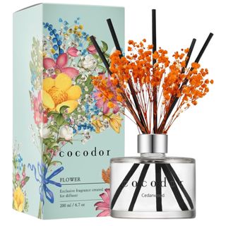 A glass cedarwood diffuser with black reeds and orange flowers next to a blue cardboard packaging box with a bright multicolored bouquet of flowers printed on it