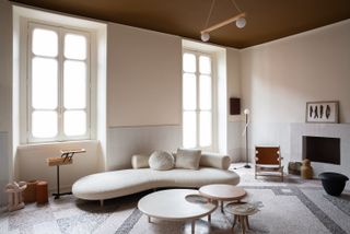 H+O Milan / A lounge room with a beige no-back sofa with cushions, two circular side tables of different heights, an open fire place, a chair, objects and floor to ceiling window and large window.