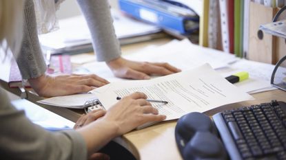 Two women look at paperwork on a desk to update it, only their hands showing.