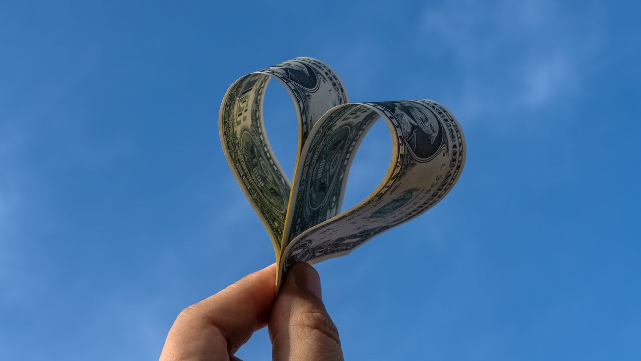 Two dollar bills shaped into a heart held against the backdrop of a blue sky.