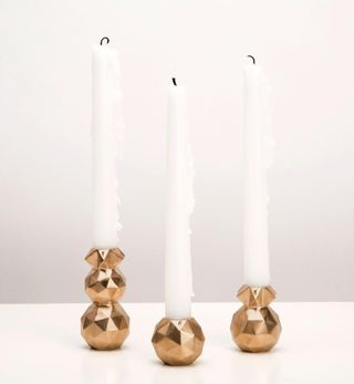 2 blownout tall white candles with geometric designed gold base. Photographed on a white surface against a white background