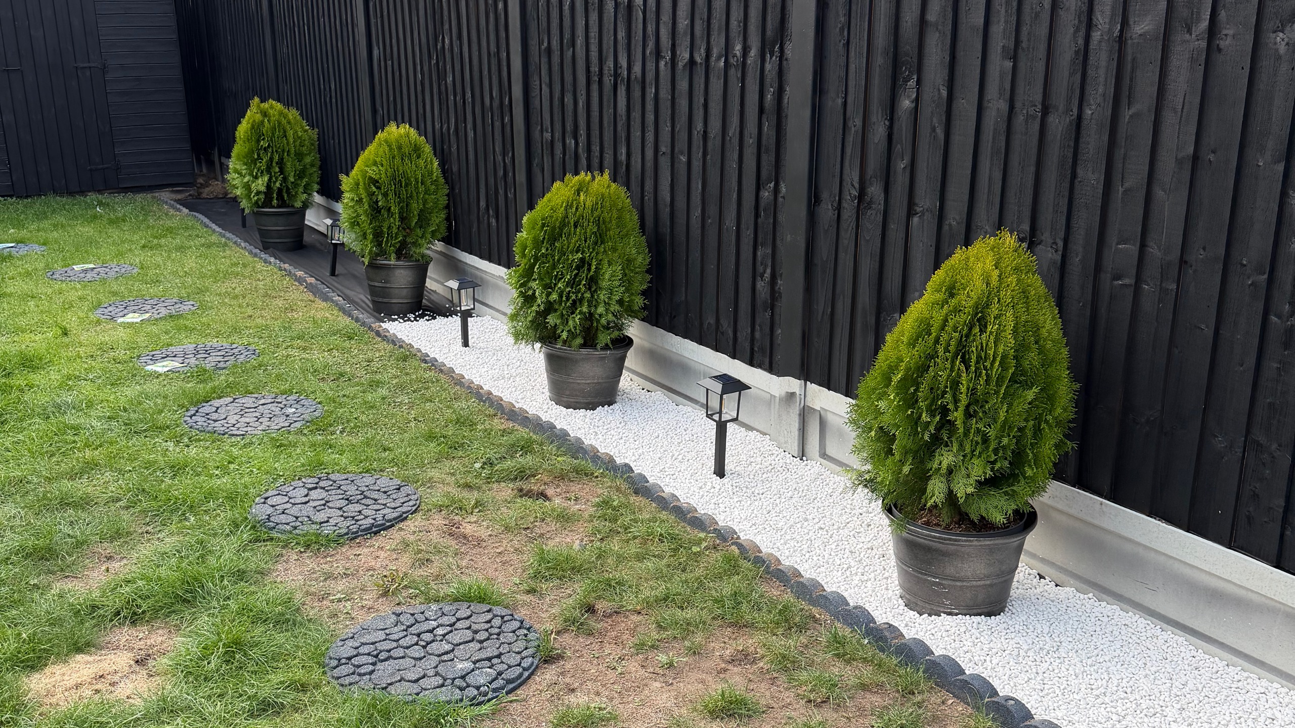 Garden with round pavers in grass, potted small trees by black fence and small lights in ground
