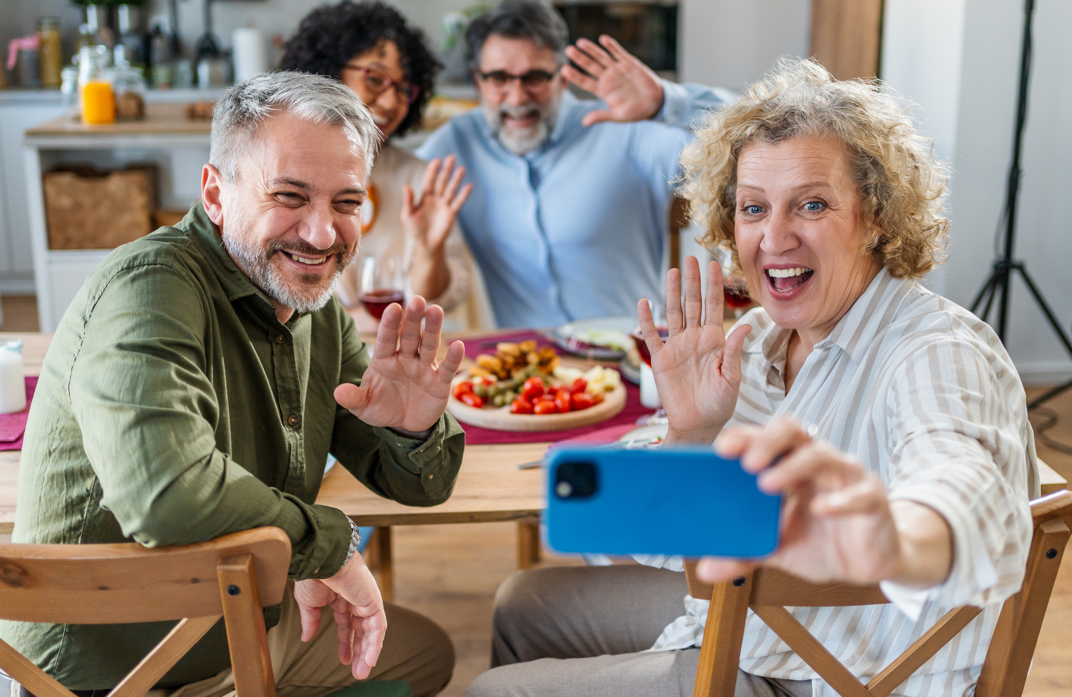 Cheerful group of diverse friends waving at smartphone camera during lunch gathering. Senior woman holding phone for selfie while others smile and gesture enthusiastically