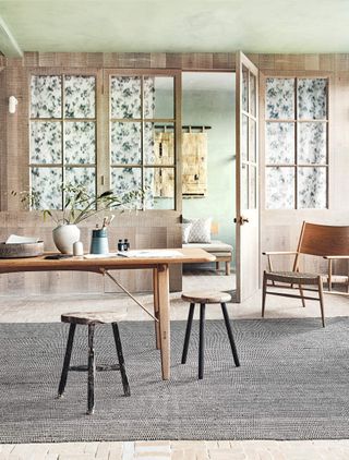 Dining room with wooden table, chair and stools on a grey rug and wooden clad and green walls.