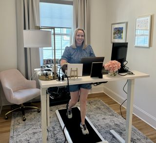 Elliott Harnell smiles at the camera as she walks on a slimline treadmill positioned under her desk. She's in her study, with a wheeled desk chair behind her and pictures framed on the wall. Her desk is filled with pictures, flowers and wires.