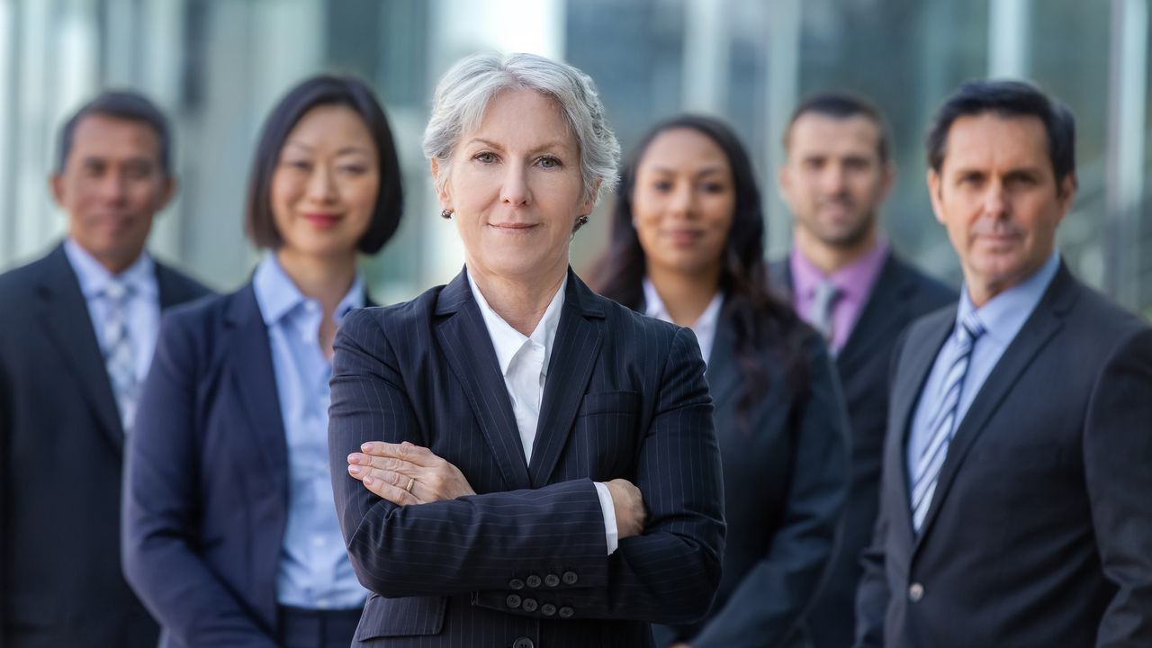 A smiling older woman who is a federal employee stands with her arms crossed with several federal employees behind her.