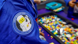 closeup of a patch on a blue shirt, which shows a spacesuited lego man smiling and waving. blurry bins of legos are in the background