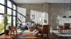 Modern living room area with brick wall, floor-to-ceiling windows and wooden floors. Pictured is a gray sofa, a leather armchair, a rattan chair, two indoor plants, a coffee table and a partially-pictured dining area to the right.