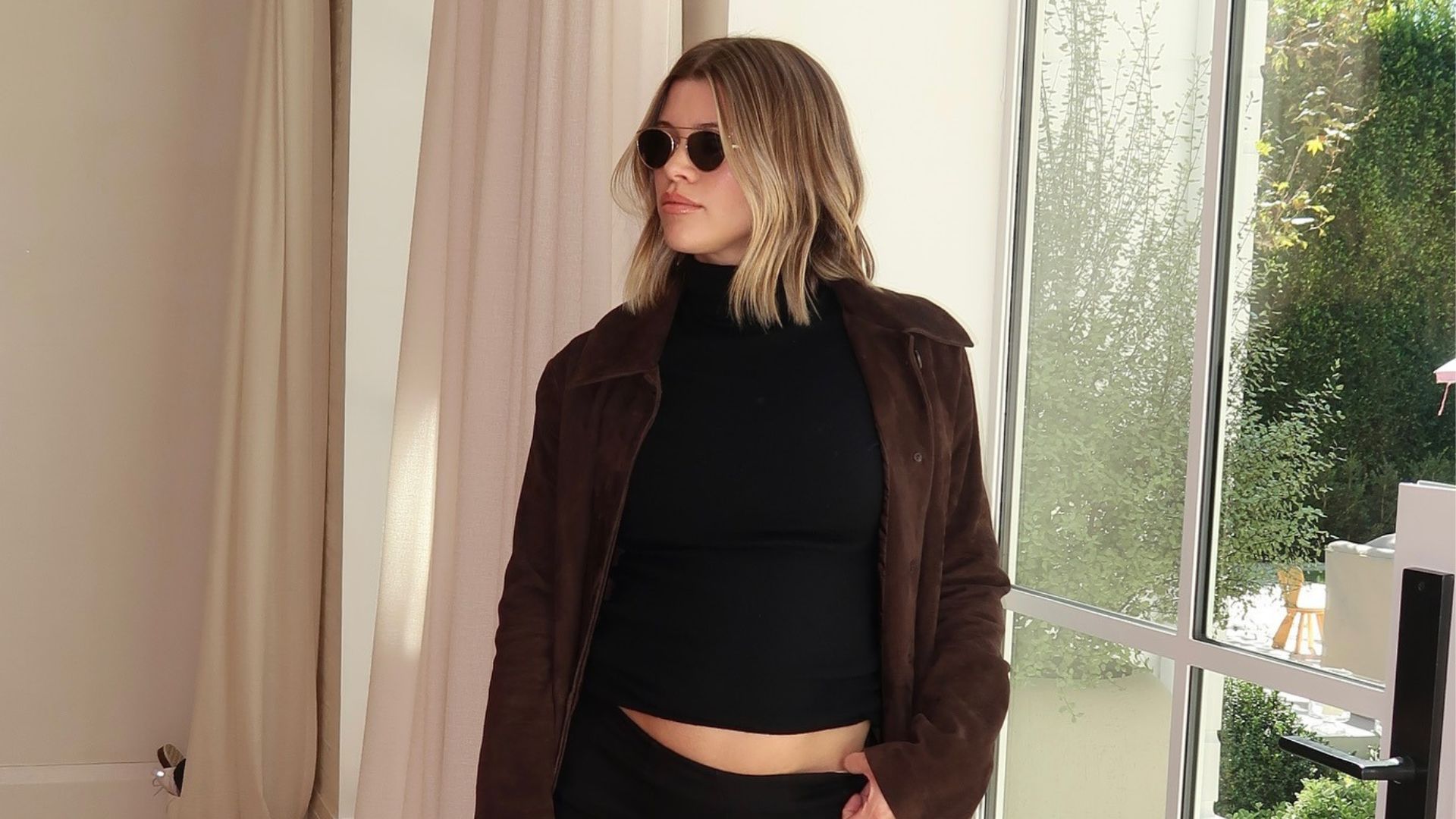 Sofia Richie Grainge poses indoors wearing a black turtleneck top with a black maxi skirt and brown suede trench coat and a black bag with chain detailing.