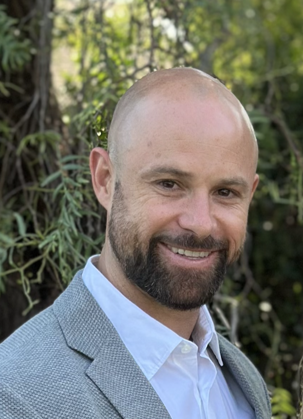 Ethan is a white man with a shaved head, brown neat beard, and is pictured wearing a button down white shirt and light gray suit jacker