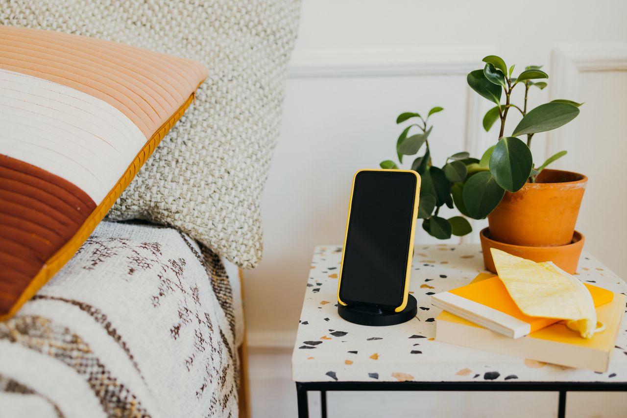 A nightstand next to a bed with an iPhone in a holder 