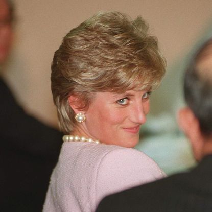 Princess Diana has short blonde hair and smirks while wearing a pink jacket and pearl jewelry