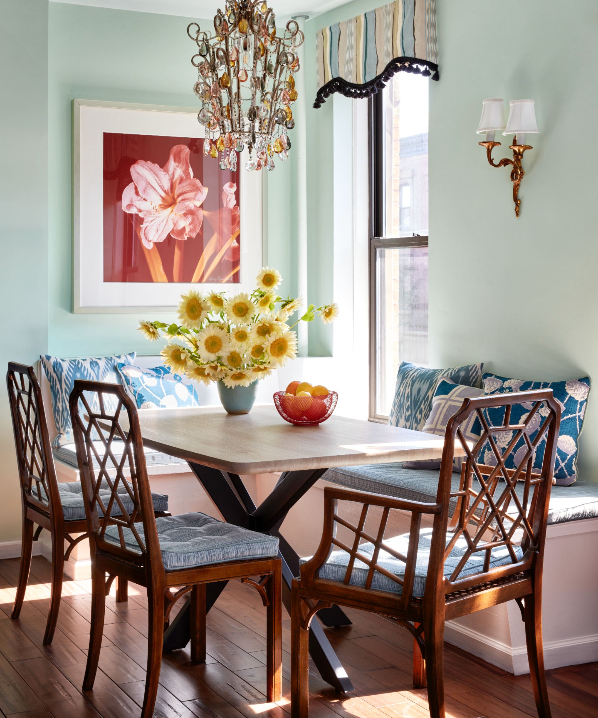 Pale blue dining nook with built-in banquette seating, a marble square table, three antique cane dining chairs, a vase of sunflowers, and an ornate chandelier lantern hanging from the ceiling.