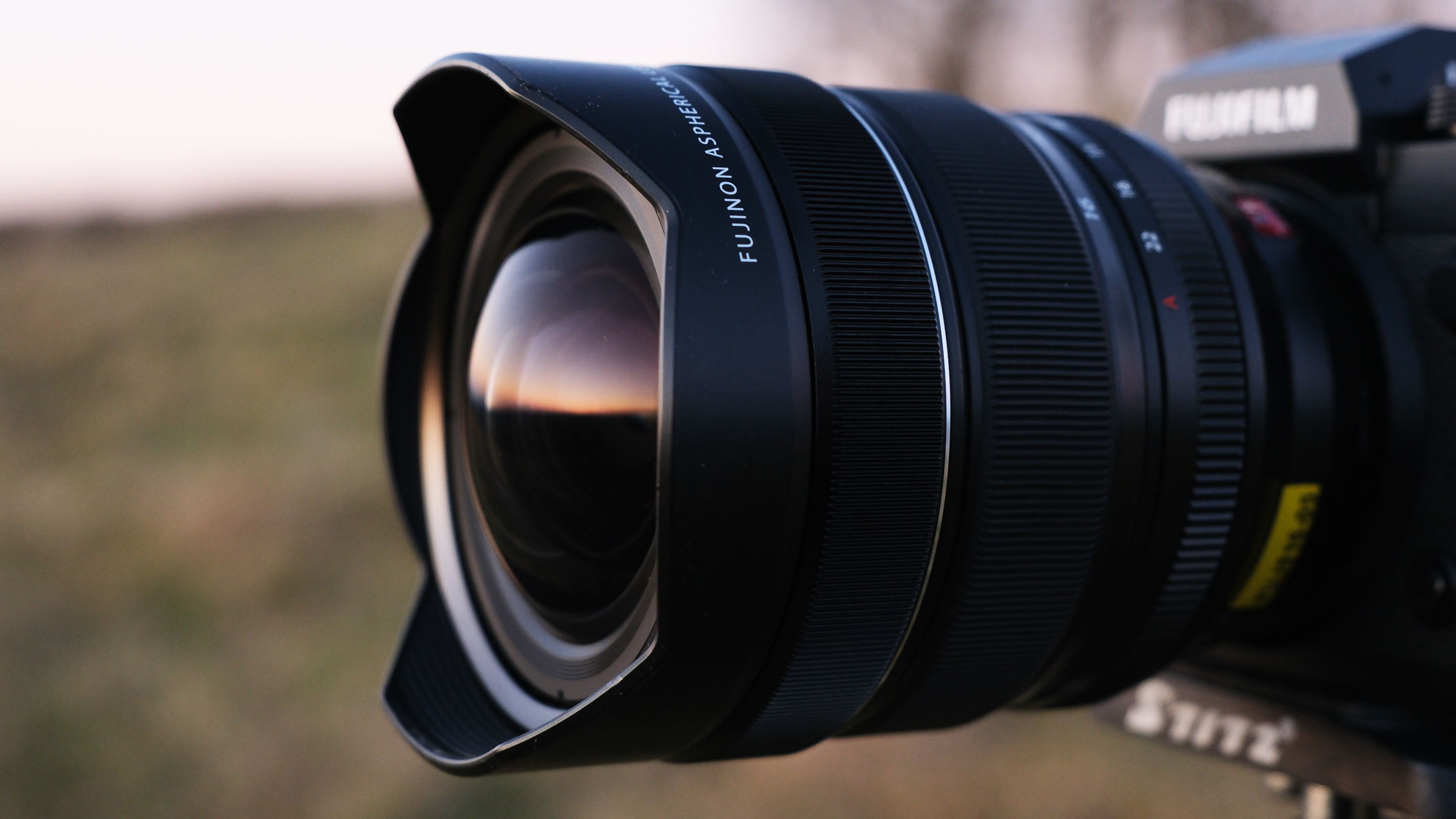 A side view of the Fujifilm X-H2 with the Fujifilm XF 8-16mm f/2.8 lens attached and a field in the background