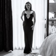 Actress Maisy Stella stands in front of a hotel window wearing a Pamella Roland black column gown with cutout and pearl detail from the Pre-Fall 25 collection. 