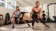 Man and woman in gym performing side lunge, with one knee bent and leaning to the side of the bent knee. The other leg is straight and they both hold dumbbells.