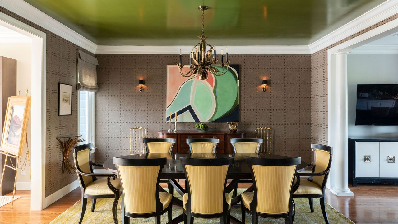 Example of ceiling paint ideas. Dining room with vivid green painted ceiling, white ceiling trim, brown patterned wallpaper, oval black dining table with eight upholstered dining chairs in yellow striped fabric, abstract wall art, two wall lights, yellow patterned rug, wooden flooring