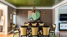 Example of ceiling paint ideas. Dining room with vivid green painted ceiling, white ceiling trim, brown patterned wallpaper, oval black dining table with eight upholstered dining chairs in yellow striped fabric, abstract wall art, two wall lights, yellow patterned rug, wooden flooring