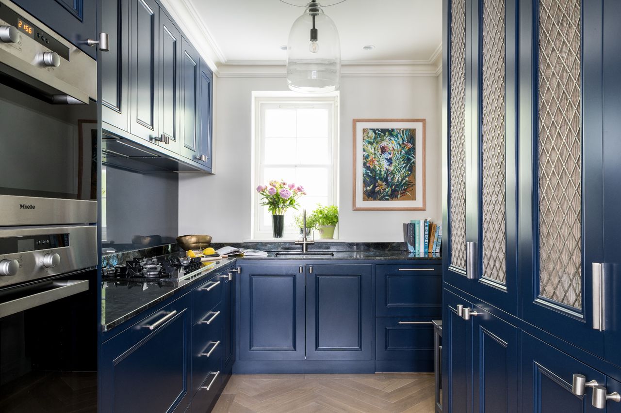 A kitchen fitted with blue cabinets, with a vase of flowers by the sink, two glass pendant lights above and light parquet floors