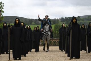 Host Alan Cumming rides a horse between rows of people in black coats and gold masks, in 'The Traitors' season 3.