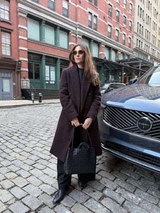 Sara Holzman is wearing a chocolate brown coat, a grey sweater, black trousers, black mules, and a brown Andiamo tote bag.